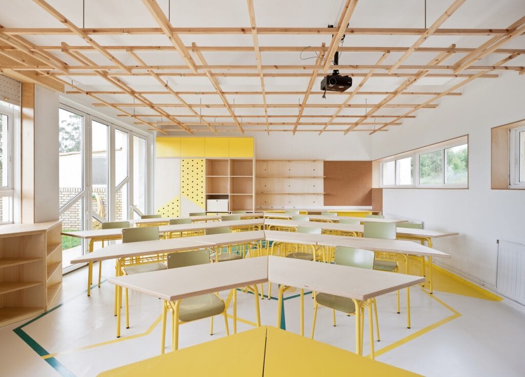 Aula transformada con nuevo mobiliario de madera, puerta acristalada y suelo con dibujos geométricos, de tonos blanco, amarillo y azul turquesa.